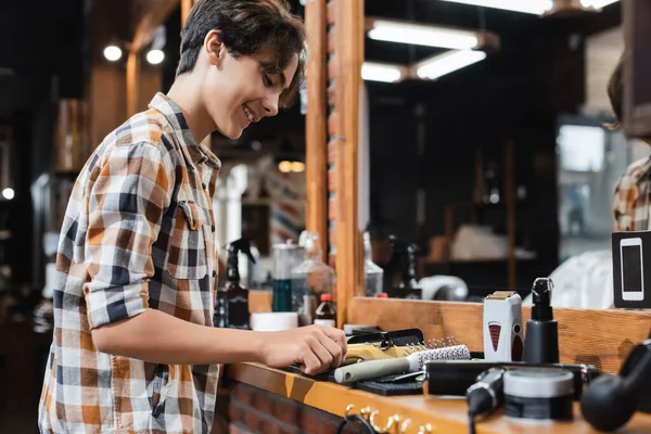 Seitenansicht eines lächelnden Teenagers, der Friseurutensilien in der Nähe eines Spiegels im Friseursalon berührt — Stockfoto