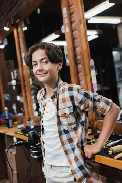 Positive teenage boy looking at camera near worktop and mirror in barbershop — Stock Photo