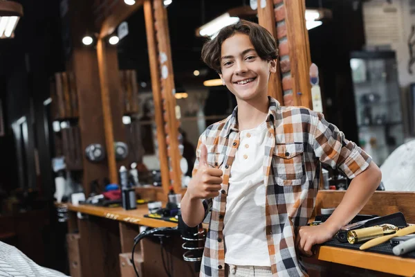 Alegre adolescente mostrando el pulgar hacia arriba cerca de herramientas de peluquería y espejo en el salón de belleza - foto de stock