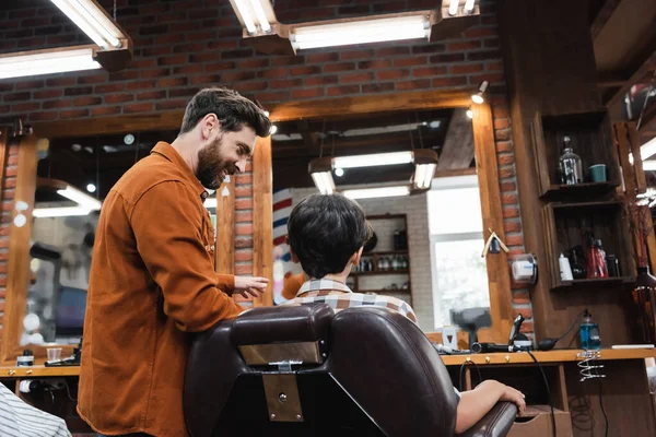 Sourire barbier parler à adolescent garçon assis dans fauteuil dans salon de coiffure — Photo de stock