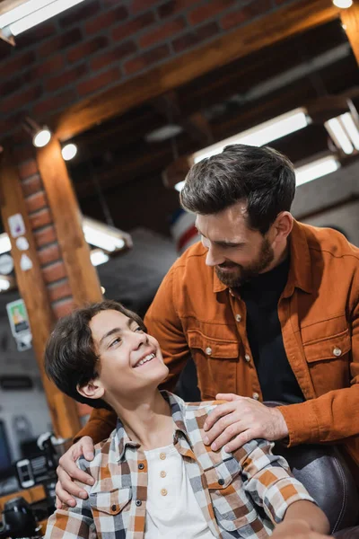 Smiling barber hugging teenager in armchair in barbershop — Stock Photo