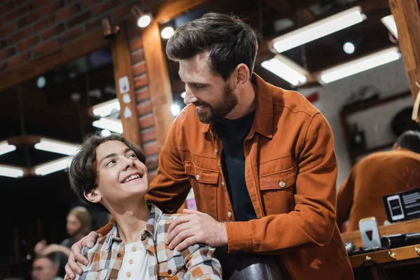Barbeiro sorridente abraçando cliente adolescente na barbearia — Fotografia de Stock