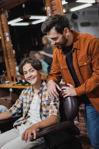 Cheerful teenage customer looking at camera while sitting in armchair near barber in barbershop — Stock Photo