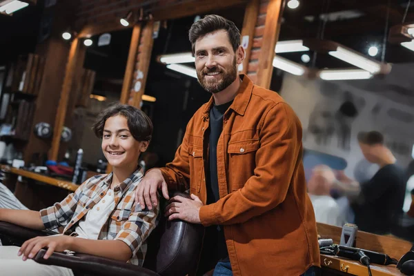 Barbeiro barbudo positivo sorrindo para a câmera perto de cliente adolescente na barbearia — Fotografia de Stock