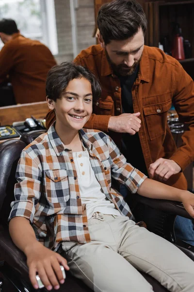 Sonriente adolescente mirando a la cámara mientras está sentado cerca de barbero en la barbería - foto de stock