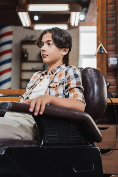Adolescente mirando hacia otro lado mientras está sentado en el sillón en la barbería - foto de stock