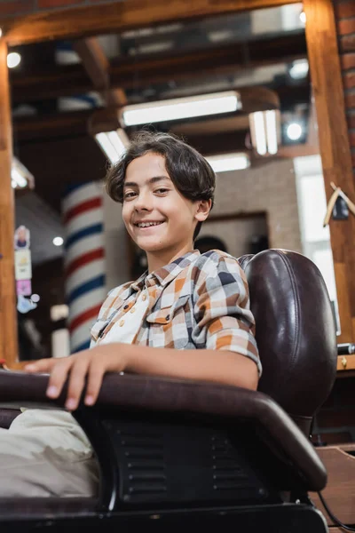 Teenage client smiling at camera while sitting in armchair in barbershop — Stock Photo