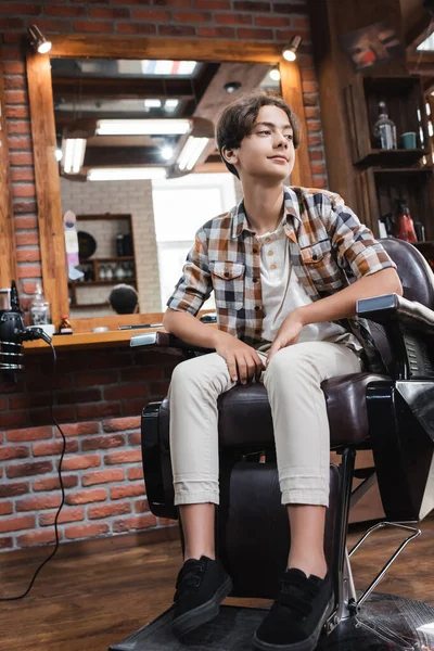 Positive teenager sitting on armchair and looking away in barbershop — Stock Photo