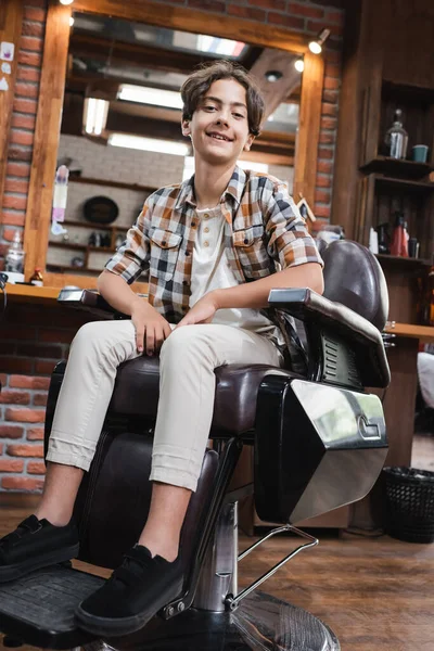 Cliente adolescente con camisa a cuadros mirando a la cámara mientras está sentado en el sillón en la barbería - foto de stock