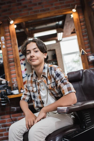 Adolescent garçon dans casual vêtements regarder caméra tandis que assis sur fauteuil dans barbershop — Photo de stock