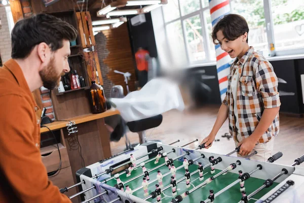 Excited teenager playing table football with blurred father in barbershop — Stock Photo