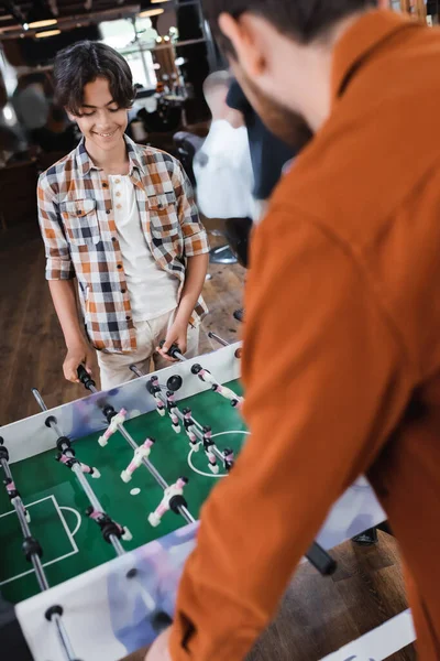 Smiling teen son playing table football with blurred father in barbershop — Stock Photo