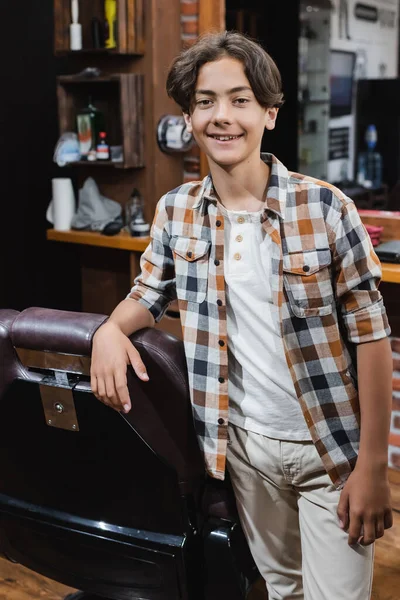 Sorridente ragazzo adolescente guardando la fotocamera vicino poltrona in barbiere — Foto stock