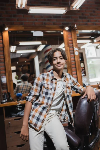 Positive teen boy looking at camera near armchair in blurred barbershop — Stock Photo