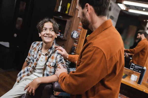 Cheerful teenager looking at blurred barber in barbershop — Stock Photo