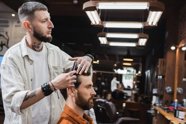 Tatuado barbeiro pentear o cabelo do homem na barbearia — Fotografia de Stock