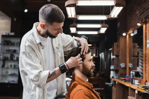 Татуйований перукар розчісує волосся усміхненого чоловіка в перукарні — стокове фото