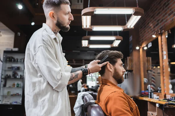 Vue latérale du coiffeur tatoué tenant peigne et pointant du doigt les cheveux du client dans le salon — Photo de stock