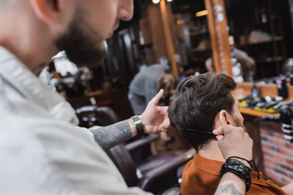 Barbeiro tatuado desfocado pentear o cabelo do homem na barbearia — Fotografia de Stock