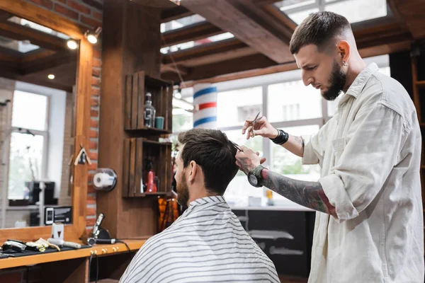 Peluquero tatuado peinando el cabello del cliente y sosteniendo tijeras en la barbería - foto de stock