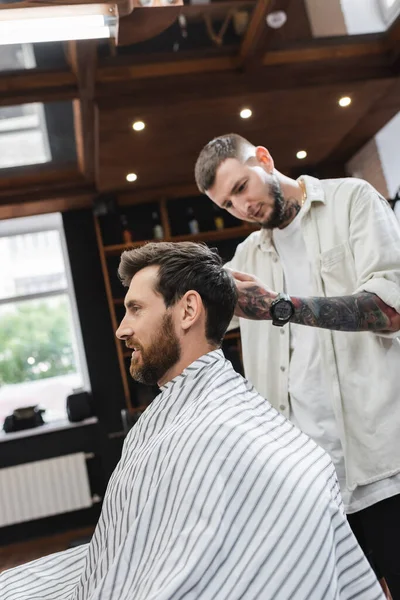 Sonriente hombre en la capa de peluquería sentado cerca de peluquero borrosa en la barbería - foto de stock