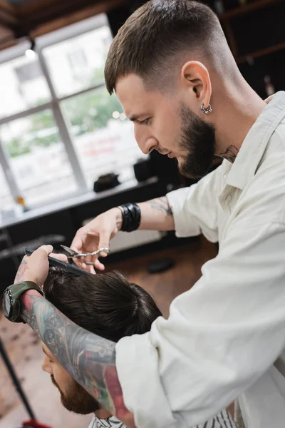 Вид збоку татуйованого перукаря, що ріже волосся клієнта в перукарні — стокове фото