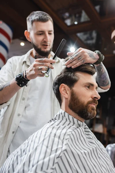 Visão de baixo ângulo de barbeiro desfocado segurando tesoura e pente ao tocar o cabelo do cliente na barbearia — Fotografia de Stock