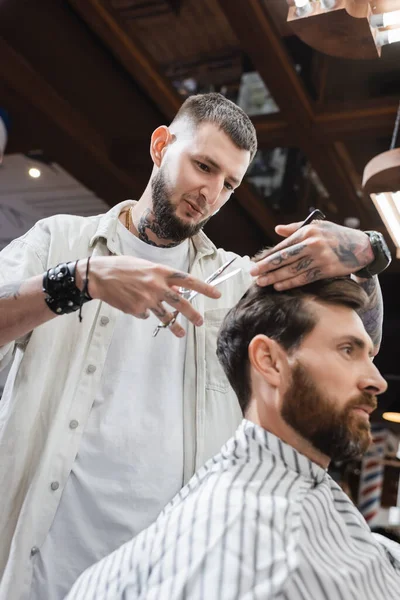 Peluquero tatuado cortando cabello de cliente barbudo en salón de belleza - foto de stock