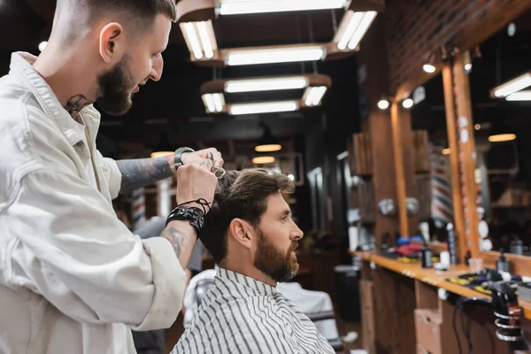 Vista lateral del peluquero tatuado cortando el cabello del cliente en la capa de peluquería en el salón de belleza - foto de stock
