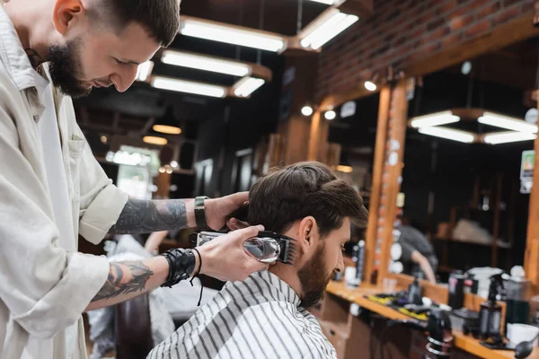 Side view of tattooed barber holding trimmer near client in barbershop — Stock Photo