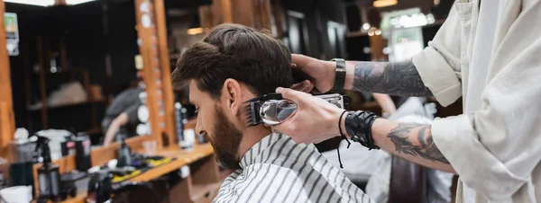 Татуйований перукар тримає тример біля бородатого клієнта в перукарні, банер — стокове фото
