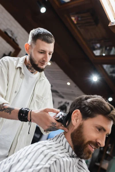 Татуированный парикмахер стрижет волосы улыбающегося клиента в салоне красоты — стоковое фото