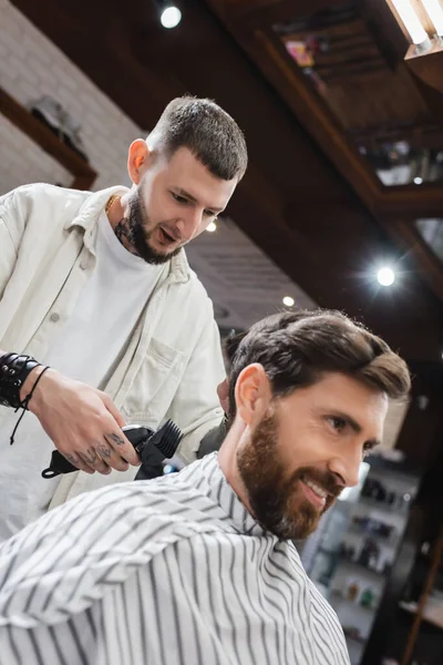 Hairstylist holding hair clipper and talking to smiling client in beauty salon — Stock Photo