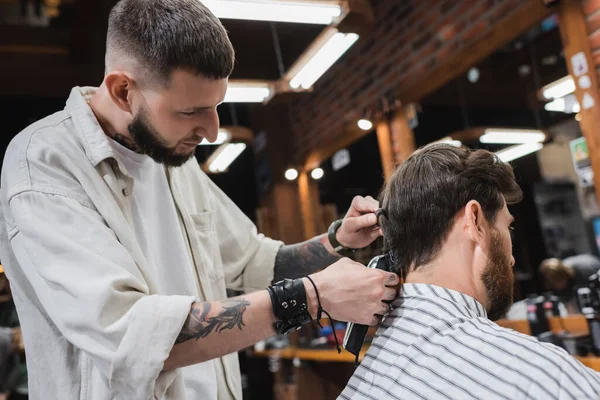 Tattooed hairstylist combing and trimming hair of brunette man in beauty salon — Stock Photo