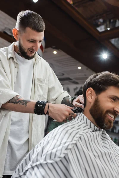 Sonriente peluquero tatuado recorte cuello de cliente barbudo en la barbería - foto de stock