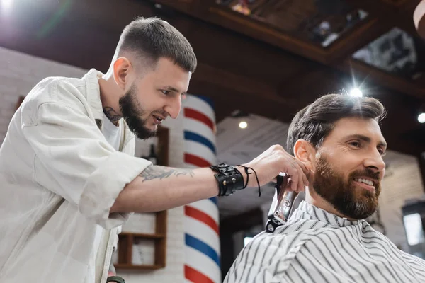 Низький кут зору татуйованого перукаря, що тримає затискач для волосся біля шиї позитивного клієнта в перукарні — стокове фото