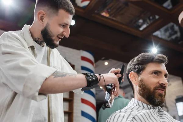 Vista de ángulo bajo del cliente sonriente sentado cerca del peluquero con trimmer y peine en la barbería - foto de stock
