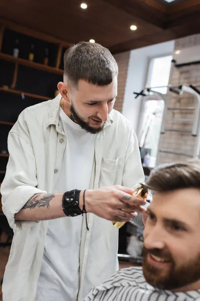 Peluquero positivo sosteniendo cortapelos cerca de cliente barbudo borroso en la barbería - foto de stock