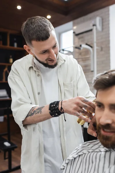 Cheerful barber trimming hair of blurred customer in hairdressing cape in barbershop — Stock Photo