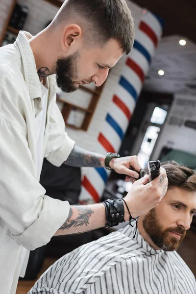 Tattooed hairstylist trimming hair of bearded man in barbershop — Stock Photo