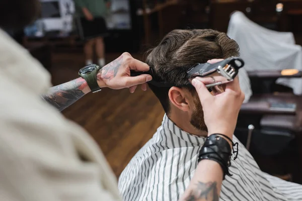 Peluquero tatuado recortando y peinando el cabello del cliente en la barbería - foto de stock