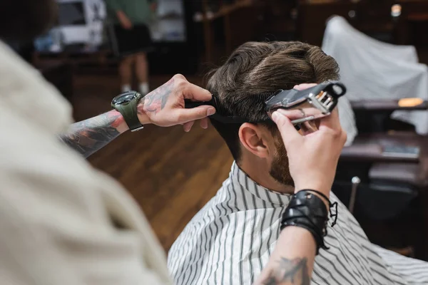 Tattooed hairstylist trimming and combing hair of customer in barbershop — Stock Photo