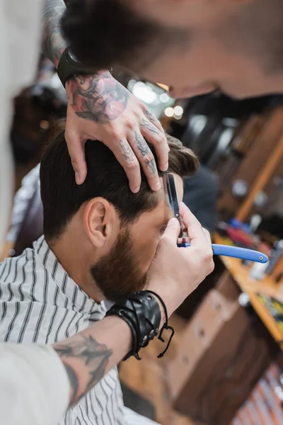 Tatuado barbeiro aparar o cabelo do cliente com navalha reta na barbearia — Stock Photo