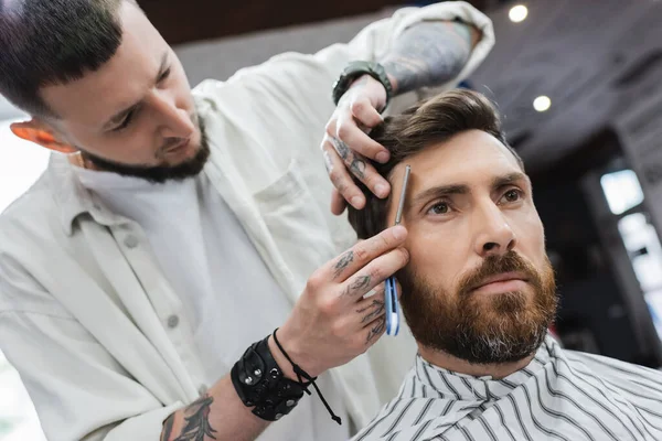 Morena barbudo hombre cerca de barbero afeitándose la frente con navaja de afeitar recta - foto de stock