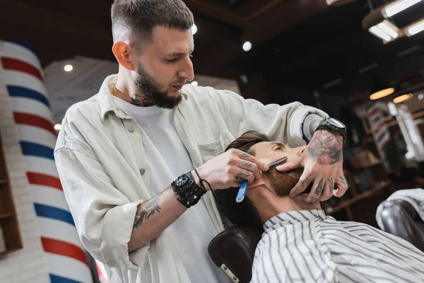 Barbeiro barbudo e tatuado barbeando cliente com navalha reta — Stock Photo