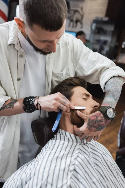 Tattooed barber in wristwatch and leather bracelet shaving client with straight razor — Stock Photo