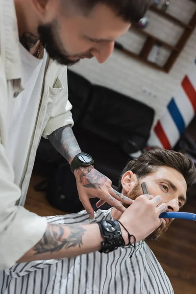 Blurred hairdresser in leather bracelet shaving cheek of man with straight leather — Stock Photo
