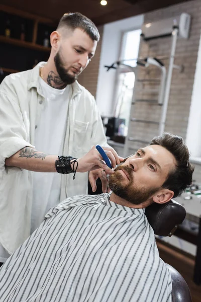 Barbudo en capa de peluquería cerca de peluquero afeitándolo con navaja de afeitar - foto de stock