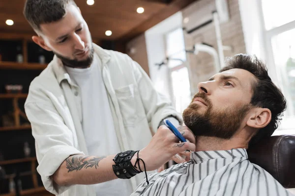 Barbeiro desfocado barbeando cliente barbudo com navalha reta na barbearia — Stock Photo