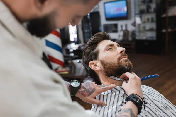 Tattooed barber shaving bearded of man with straight razor in barbershop — Stock Photo